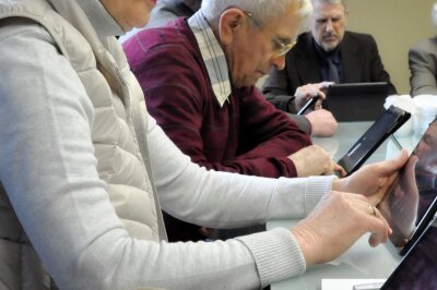 Nuotraukoje pavaizduoti trys asmenys. Jie naudojasi planšetiniais kompiuteriais. Pagrindiniame plane yra vyresnio amžiaus asmuo raudonu megztuku ir dėvintis akinius.
